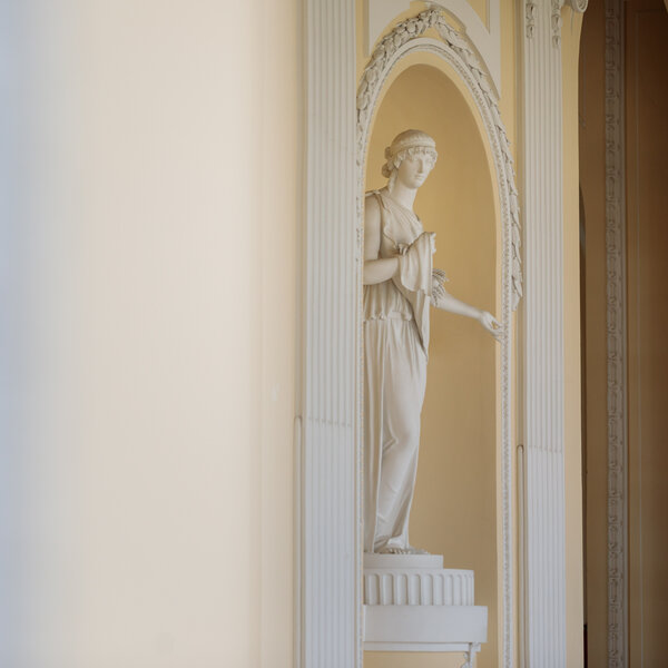 Foto: Eine weiße Statue steht in einer dekorativen Nische mit verzierten Säulen in einem Innenraum. Der Hintergrund ist hell und neutral gehalten.