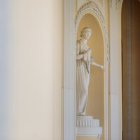 Foto: Eine weiße Statue steht in einer dekorativen Nische mit verzierten Säulen in einem Innenraum. Der Hintergrund ist hell und neutral gehalten.