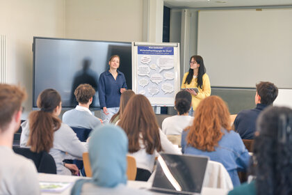 Foto: Zwei Personen präsentieren vor einer Gruppe von Studierenden. Ein Flipchart zeigt den Text 'Was bedeutet Wirtschaftspädagogik für Dich?'.