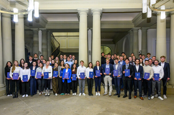 Foto: Eine Gruppe von Personen steht in einem Innenraum mit Säulen, alle halten blaue Zertifikatsmappen. Die Personen sind in formeller Kleidung und stehen in Reihen.