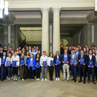 Foto: Eine Gruppe von Personen steht in einem Innenraum mit Säulen, alle halten blaue Zertifikatsmappen. Die Personen sind in formeller Kleidung und stehen in Reihen.